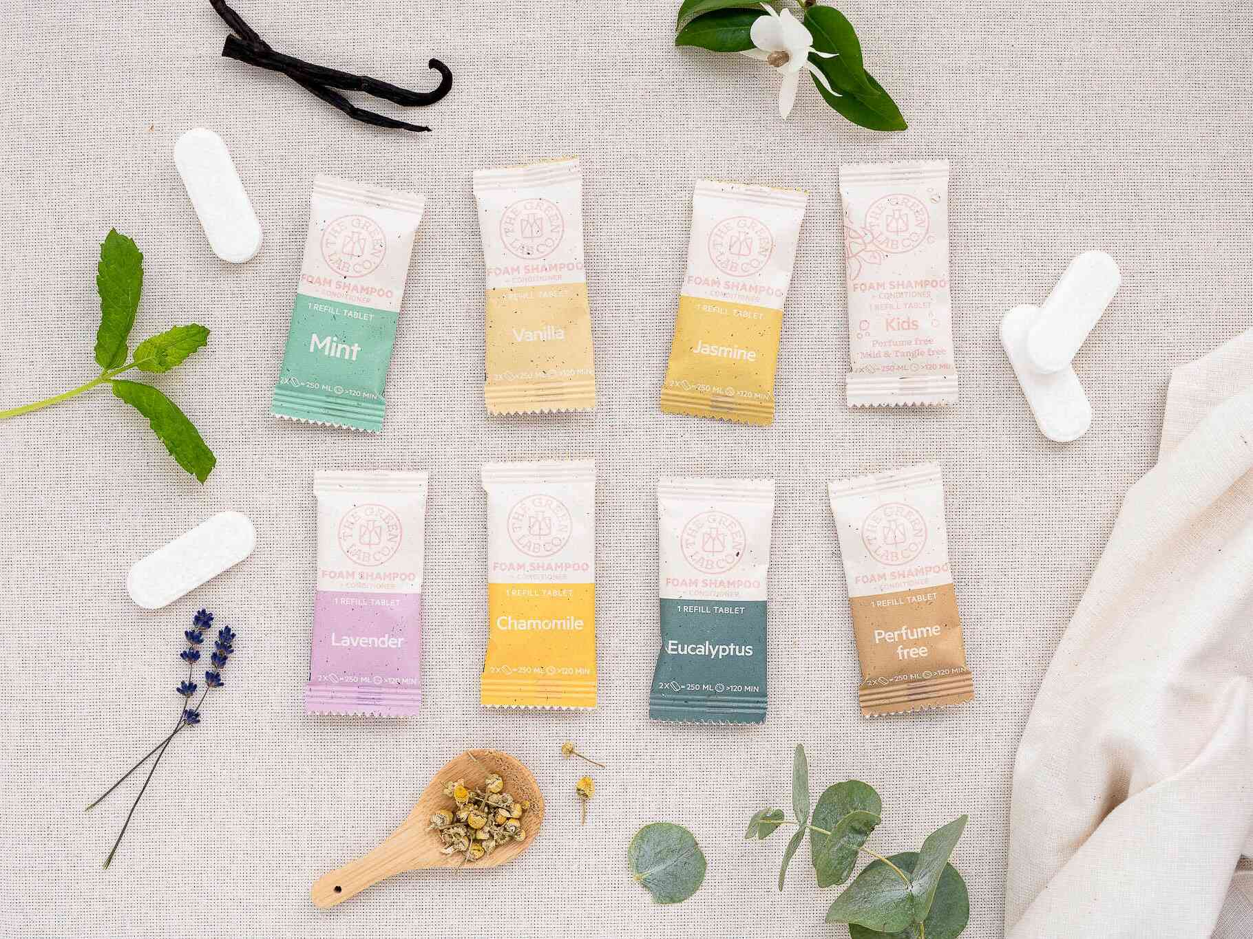 Flat lay of The Green Lab Co. foam shampoo refill tablets—Mint, Vanilla, Jasmine, Kids, Lavender, Chamomile, Eucalyptus, and Perfume Free—surrounded by botanicals on natural fabric.